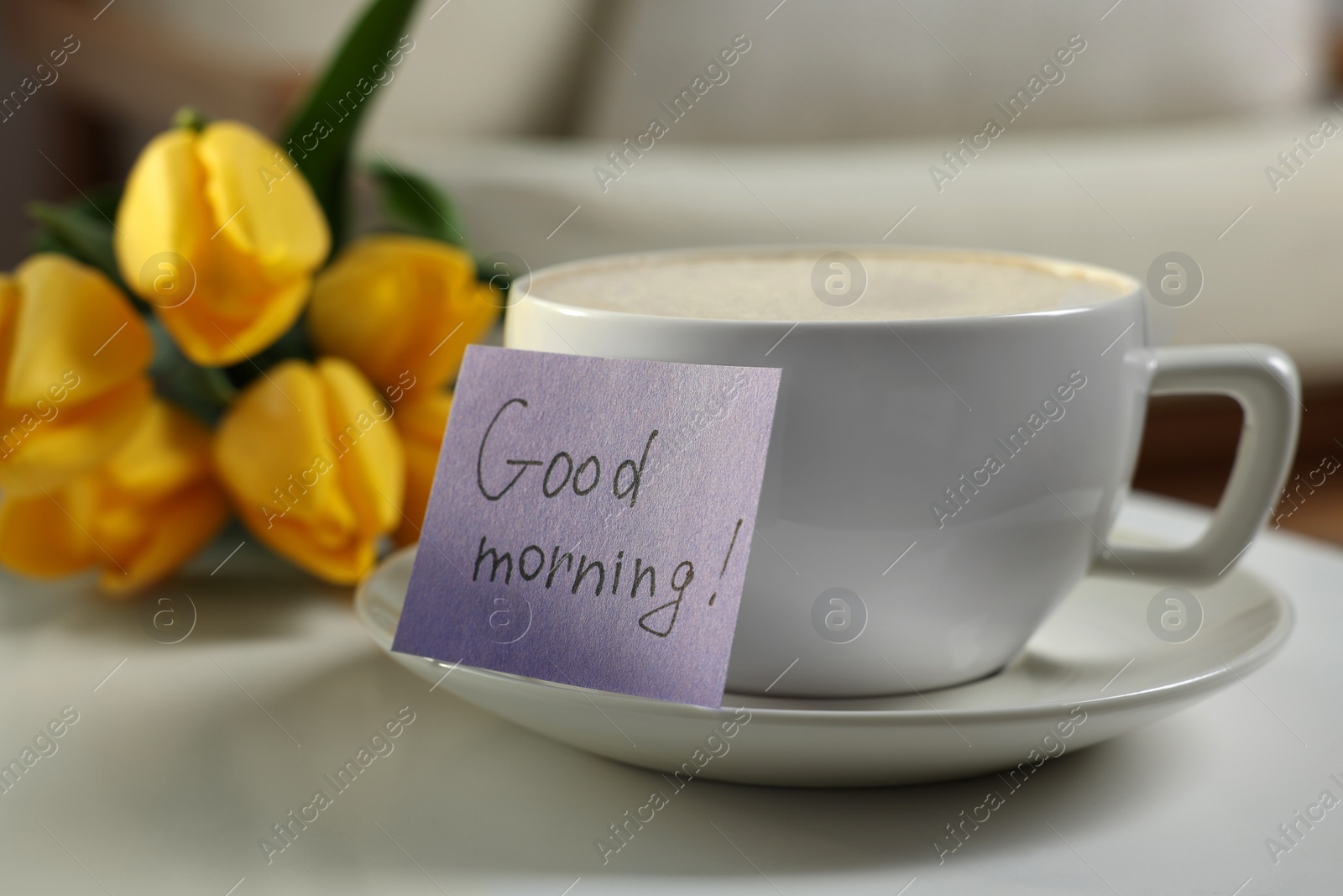 Photo of Cup of coffee, flowers and card with phrase GOOD MORNING! on white table indoors, closeup