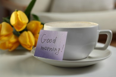 Photo of Cup of coffee, flowers and card with phrase GOOD MORNING! on white table indoors, closeup