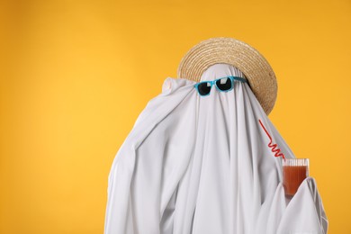 Person in ghost costume, sunglasses and straw hat holding glass of drink on yellow background, space for text