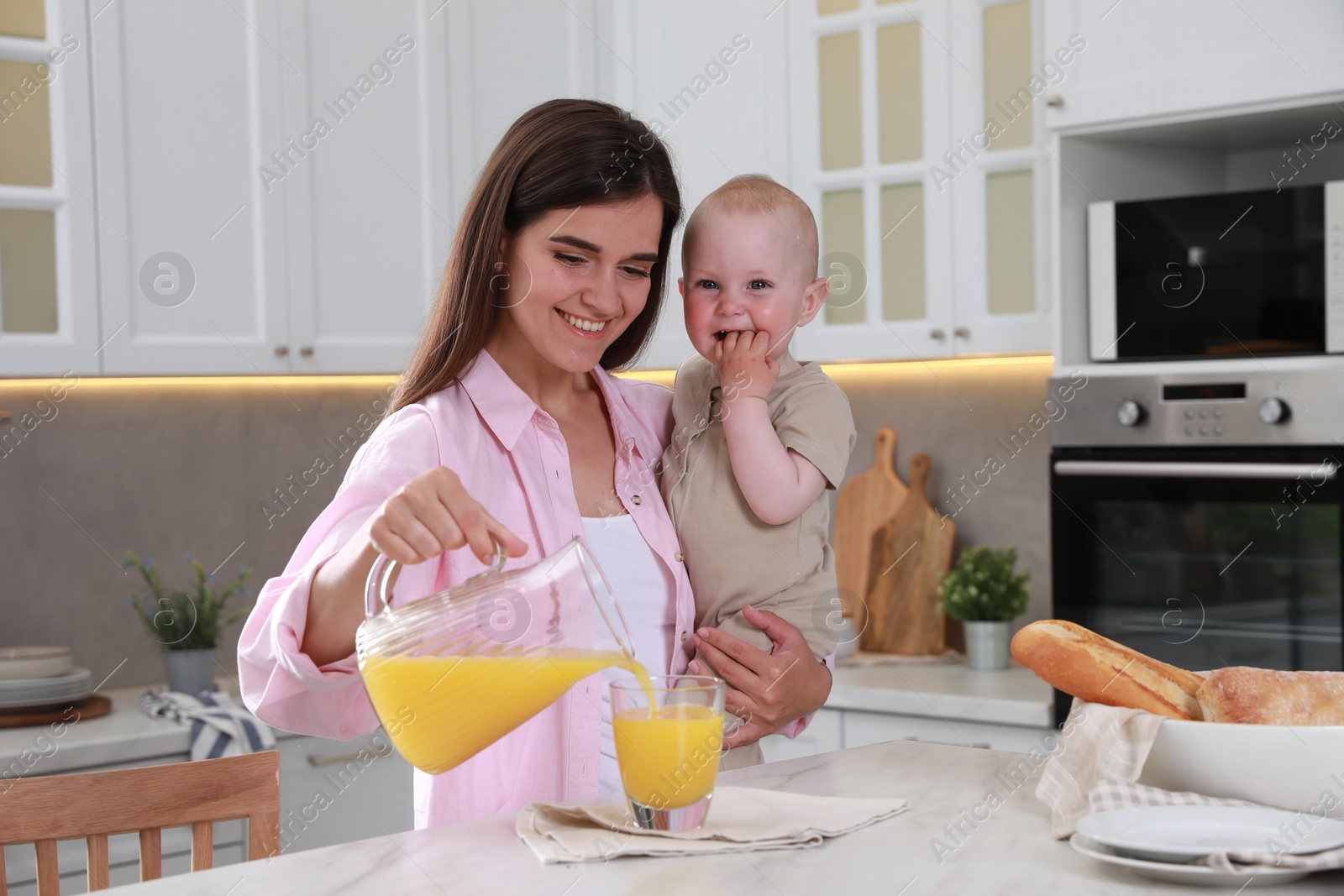 Photo of Happy young woman holding her cute little baby while pouring juice into glass in kitchen