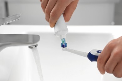 Photo of Man squeezing toothpaste from tube onto electric toothbrush above sink, closeup