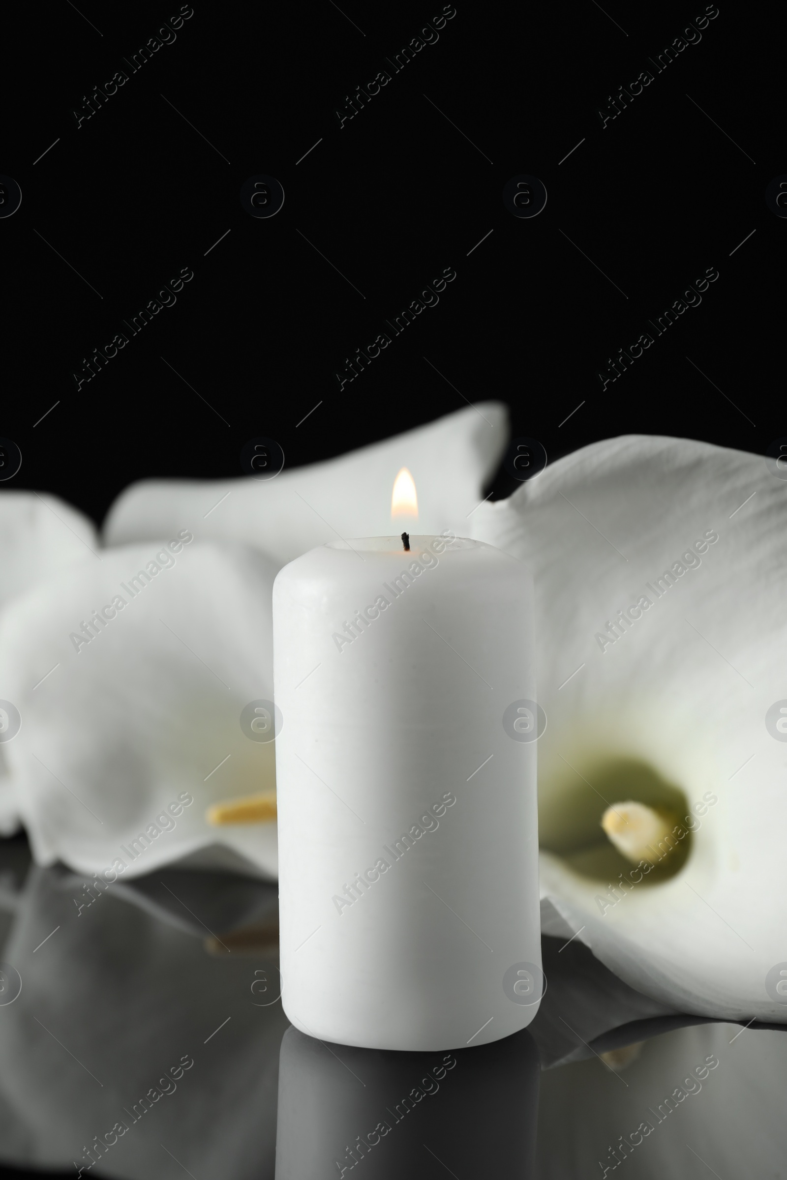 Photo of Burning candle and white calla lily flowers on black mirror surface in darkness, closeup with space for text. Funeral symbol