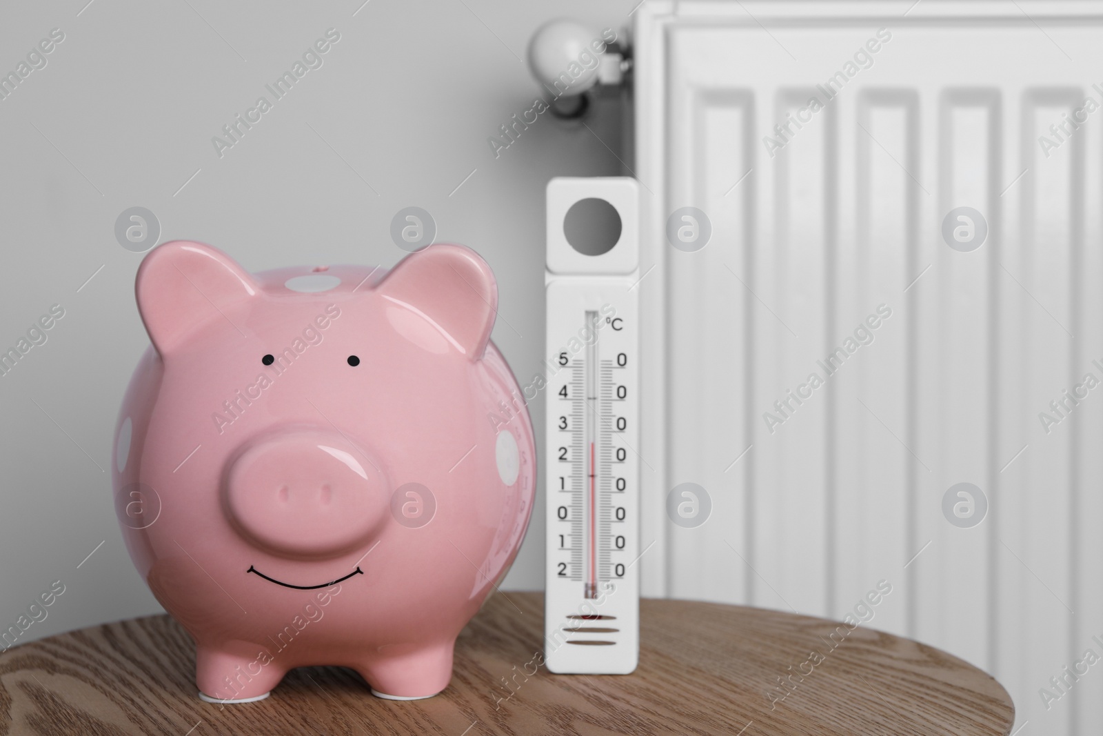 Photo of Piggy bank with thermometer on wooden table near heating radiator. Space for text