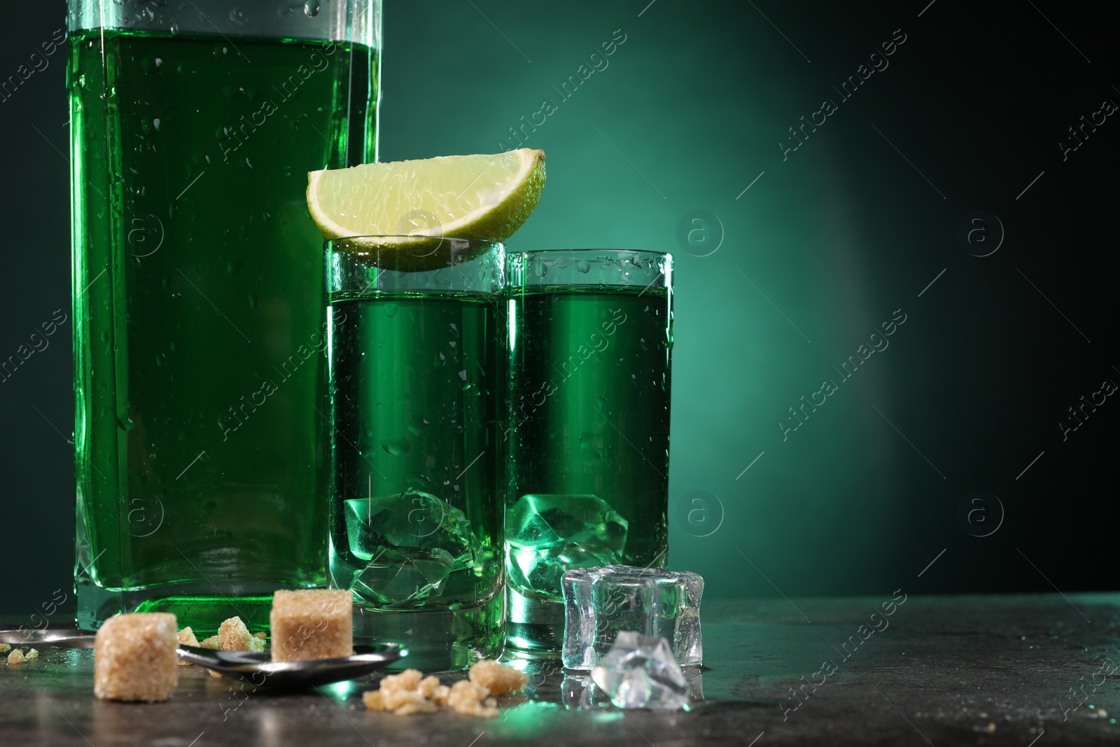 Photo of Absinthe in shot glasses with ice cubes, lime, brown sugar and spoon on gray table against green background, space for text. Alcoholic drink