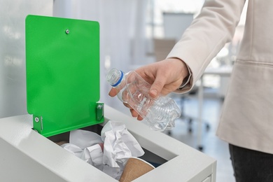 Photo of Woman putting used plastic bottle into trash bin in modern office, closeup. Waste recycling