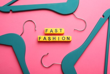 Photo of Yellow plastic cubes with phrase Fast Fashion and hangers on pink background, flat lay