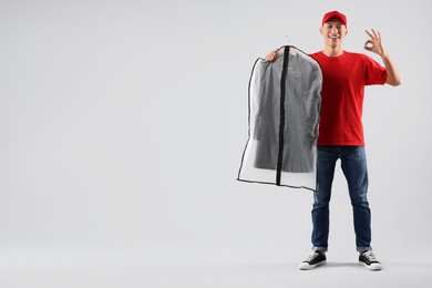 Photo of Dry-cleaning delivery. Happy courier holding garment cover with clothes and showing OK gesture on light grey background, space for text