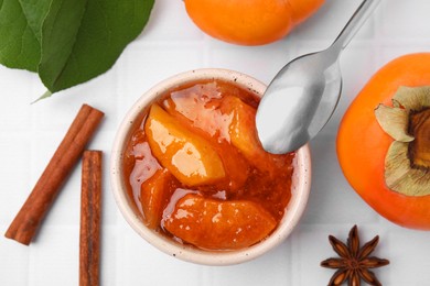 Photo of Bowl of tasty persimmon jam and ingredients on white tiled table, flat lay