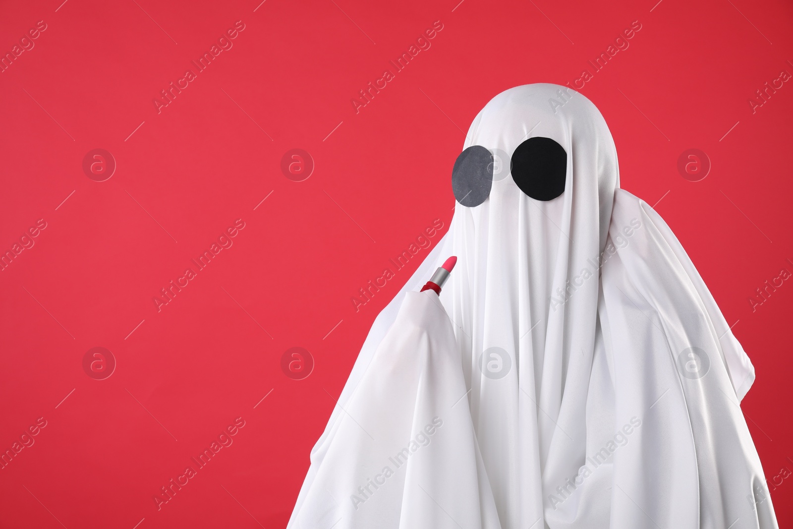 Photo of Fashionable ghost. Woman covered with white sheet applying lipstick on red background, space for text