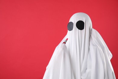 Fashionable ghost. Woman covered with white sheet applying lipstick on red background, space for text