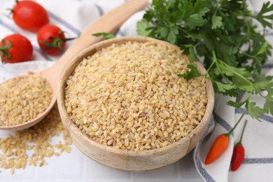 Photo of Raw bulgur in bowl, spoon, vegetables and parsley on table, closeup