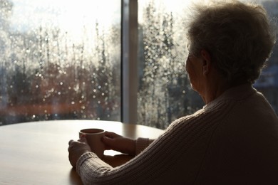 Elderly woman with cup of drink near window indoors on rainy day, space for text. Loneliness concept