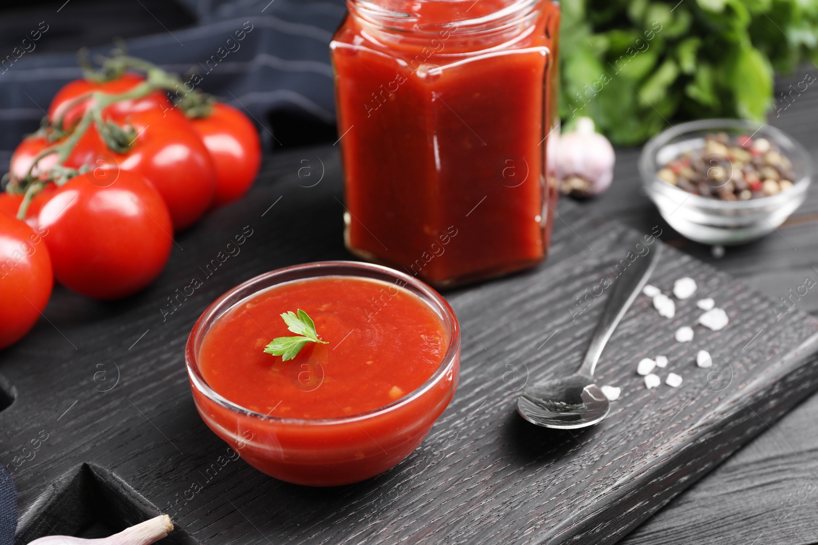 Photo of Delicious ketchup, spoon and salt on black wooden table, closeup. Tomato sauce