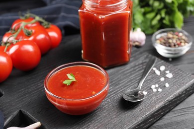 Photo of Delicious ketchup, spoon and salt on black wooden table, closeup. Tomato sauce