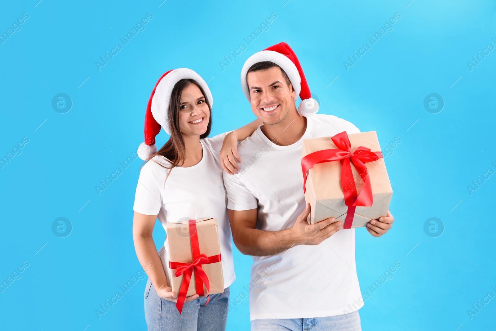 Photo of Beautiful happy couple in Santa hats holding Christmas gifts on light blue background