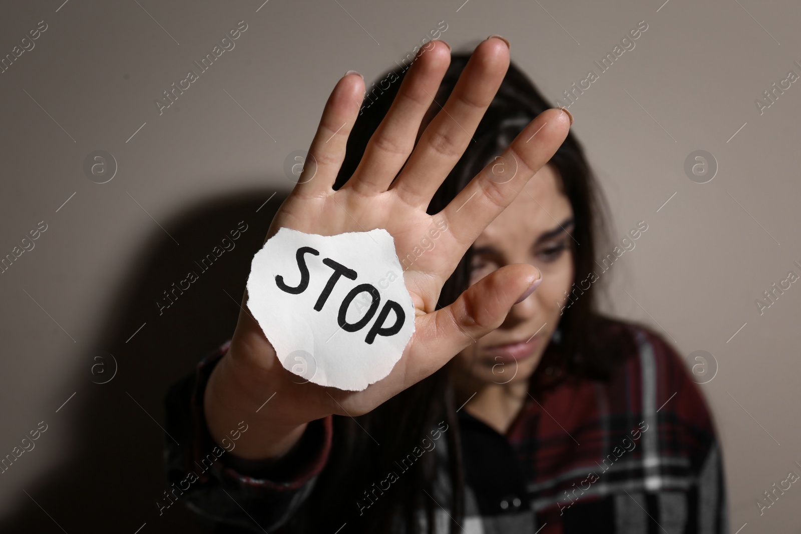 Photo of Abused young woman with sign STOP near beige wall, focus on hand. Domestic violence concept