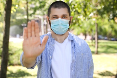 Photo of Man in protective face mask showing stop gesture in park. Prevent spreading of coronavirus