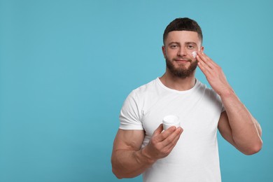 Photo of Handsome man applying cream onto his face on light blue background, space for text