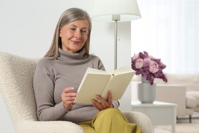 Beautiful senior woman reading book in armchair at home