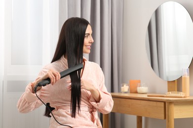 Photo of Beautiful happy woman using hair iron near mirror in room