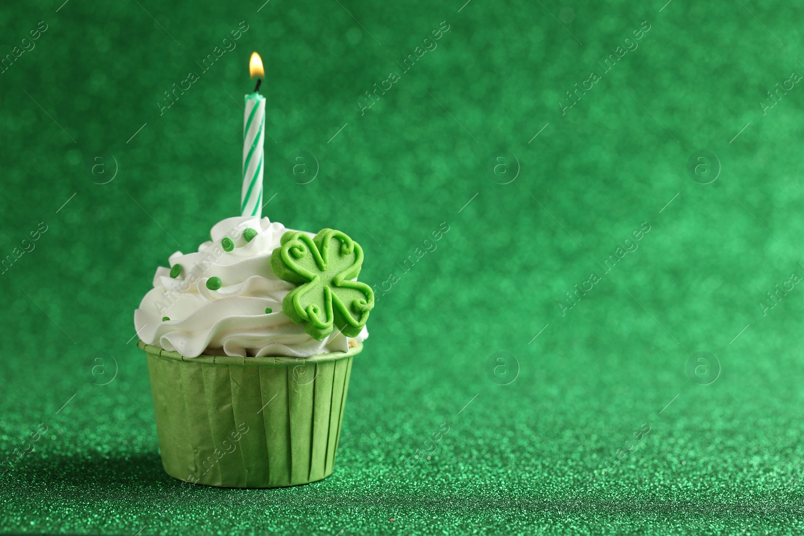 Photo of St. Patrick's day party. Tasty cupcake with clover leaf topper and burning candle on shiny green background. Space for text