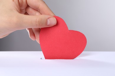 Photo of Woman putting red heart into slot of donation box against grey background, closeup