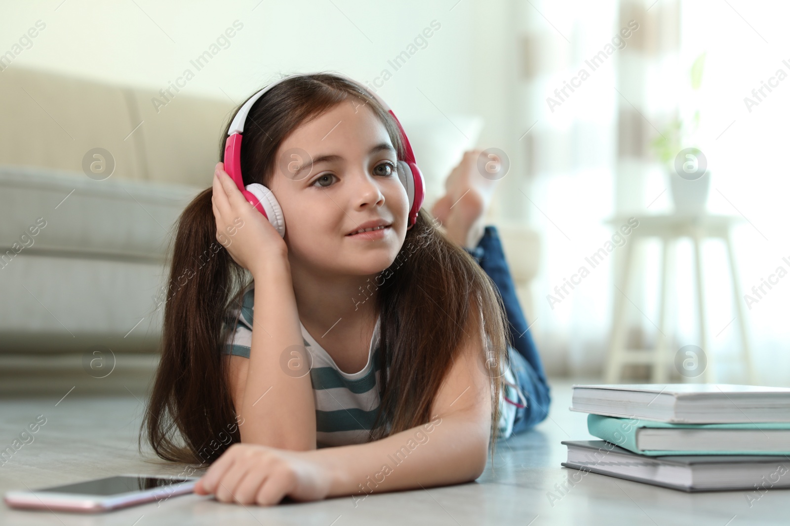 Photo of Cute little girl with headphones and smartphone listening to audiobook at home