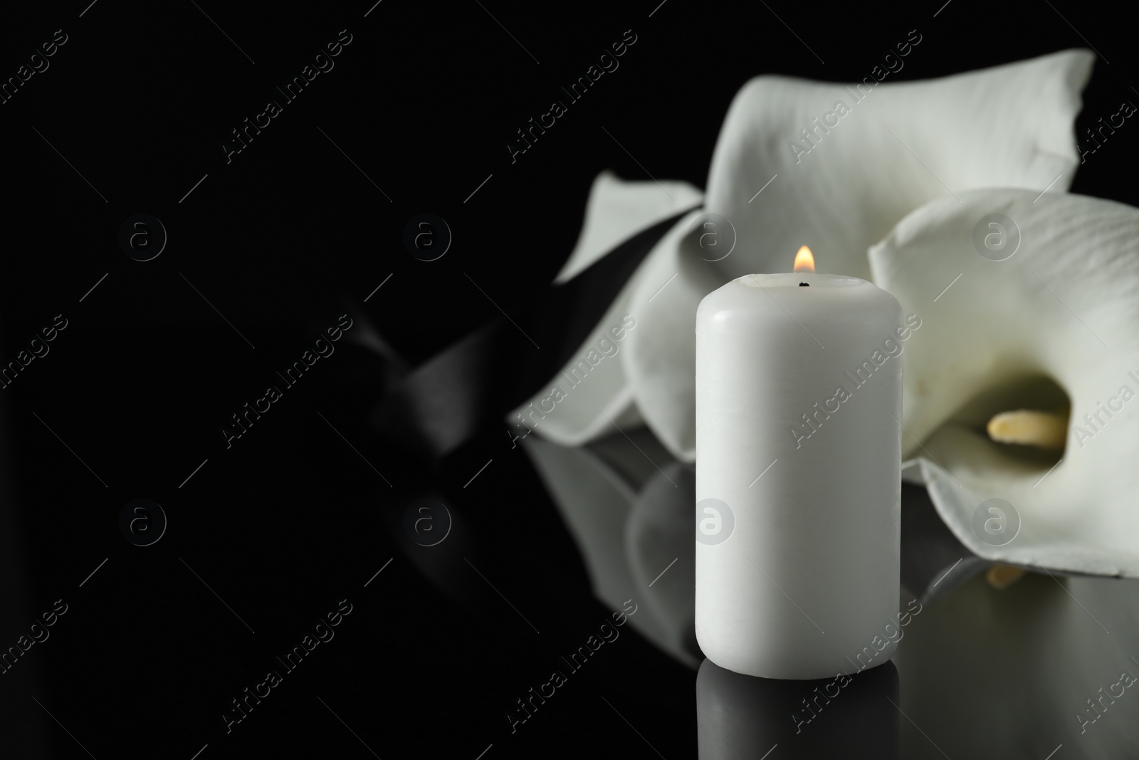 Photo of Burning candle and white calla lily flowers on black mirror surface in darkness, closeup with space for text. Funeral symbol