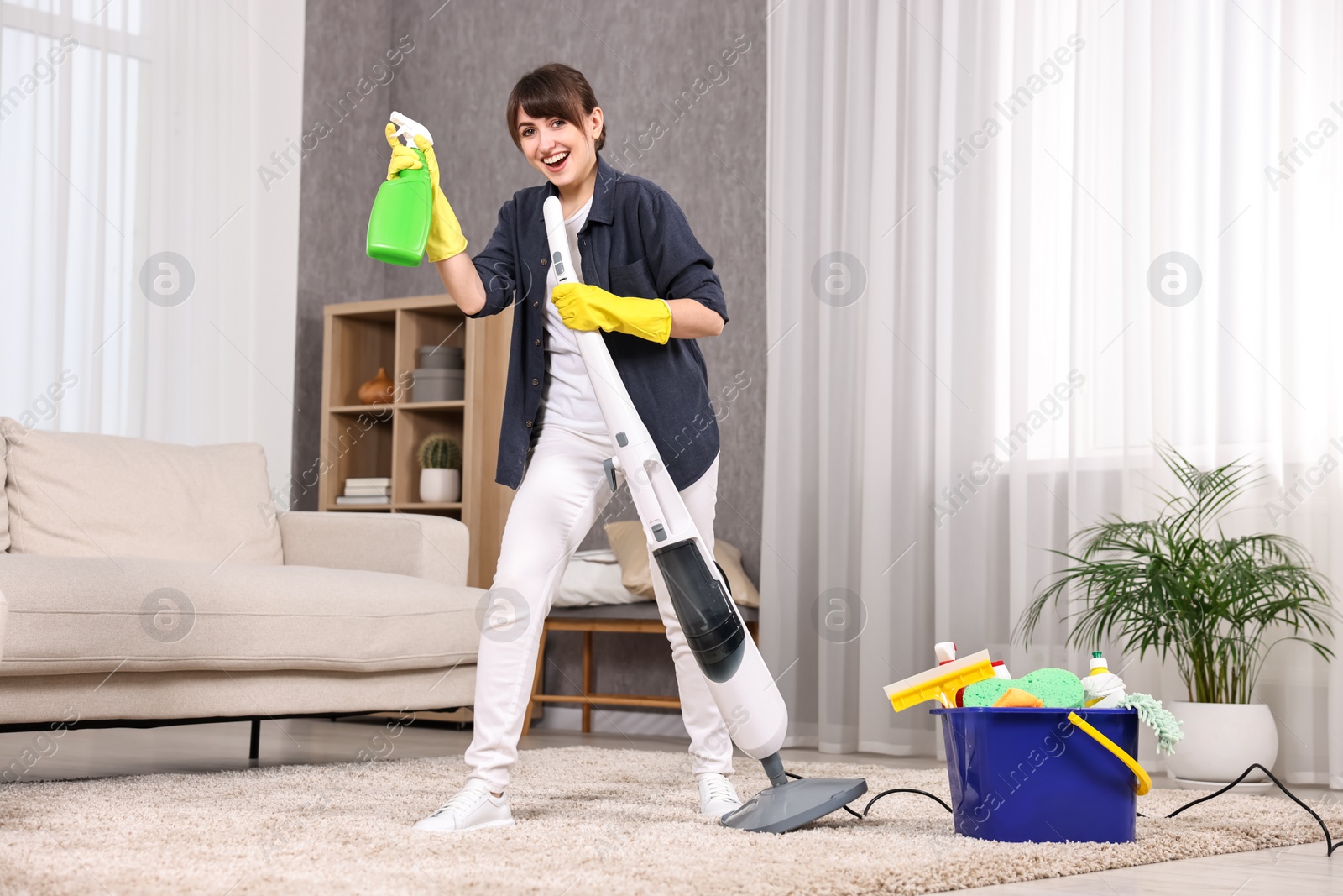 Photo of Happy young housewife with spray bottle having fun while cleaning carpet at home