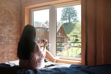 Young woman sitting on bed and enjoying view from window. Peaceful morning