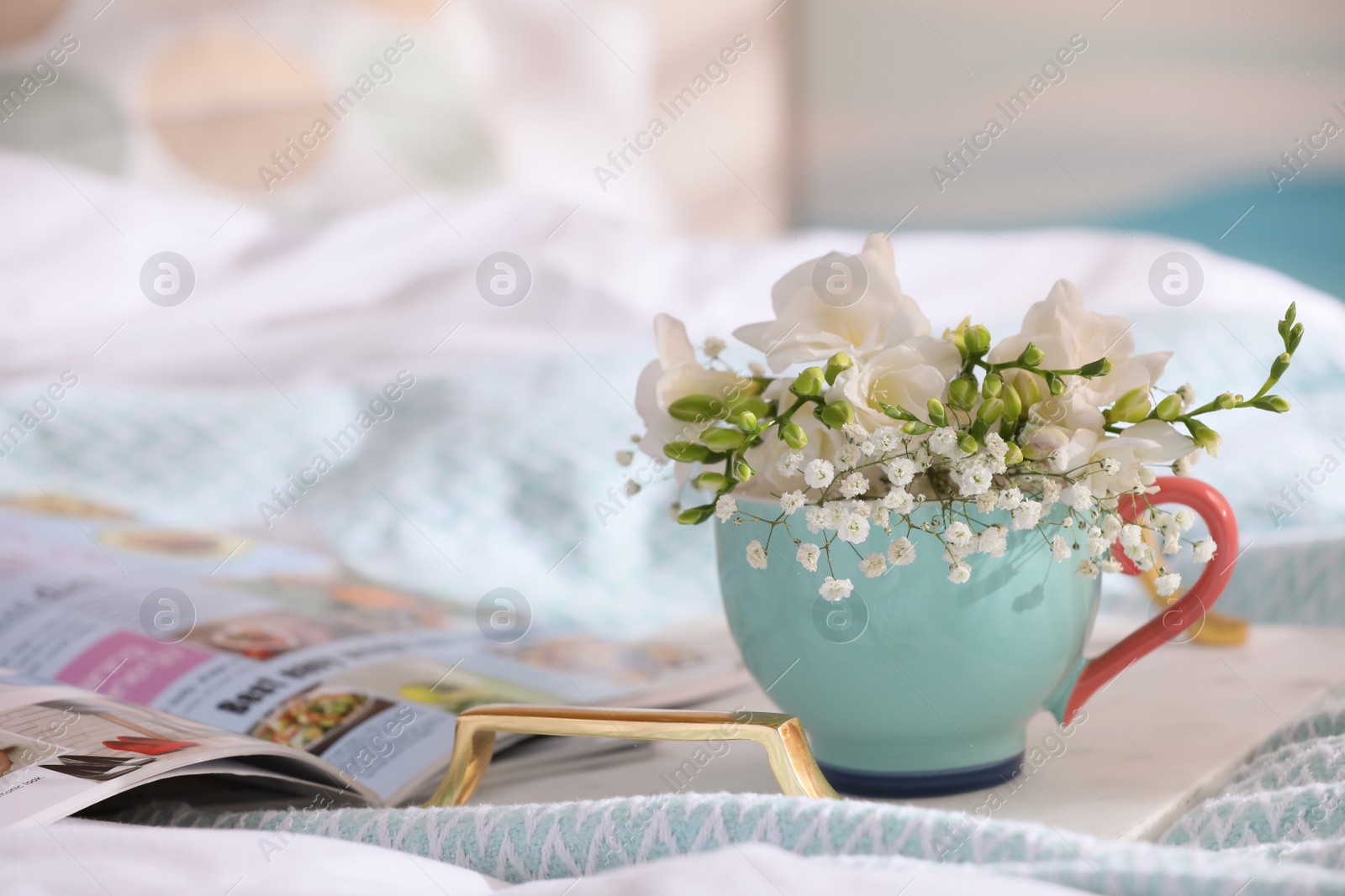 Photo of Beautiful bright flowers in cup, open magazine and tray on bed. Space for text