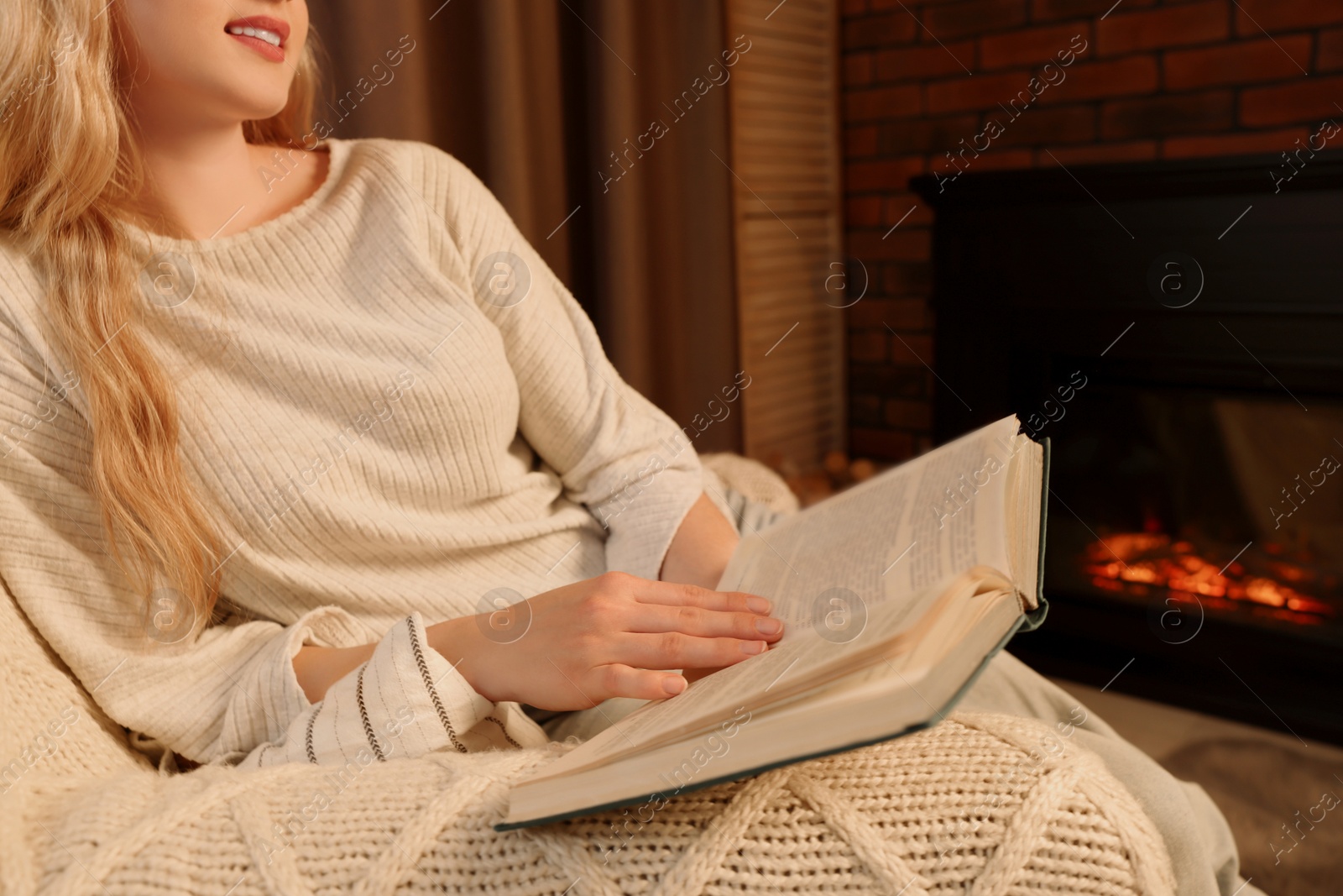 Photo of Beautiful young woman reading book near fireplace in room, closeup