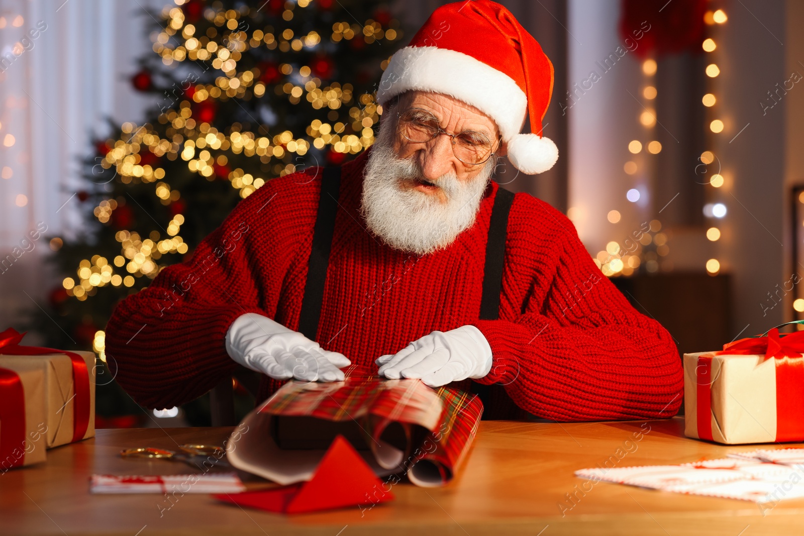 Photo of Santa Claus wrapping gift at his workplace in room decorated for Christmas