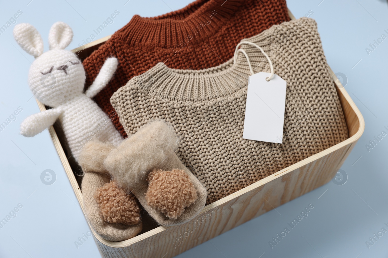 Photo of Different baby accessories, knitted sweaters and blank card in box on light blue background