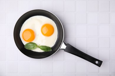 Photo of Tasty fried eggs with basil in pan on white tiled table, top view. Space for text