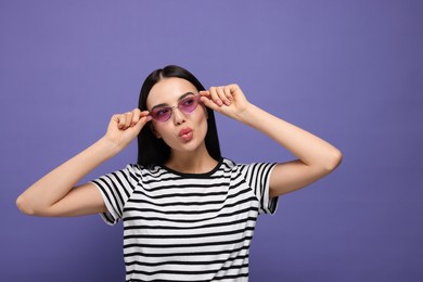 Photo of Beautiful young woman in stylish sunglasses blowing kiss on purple background