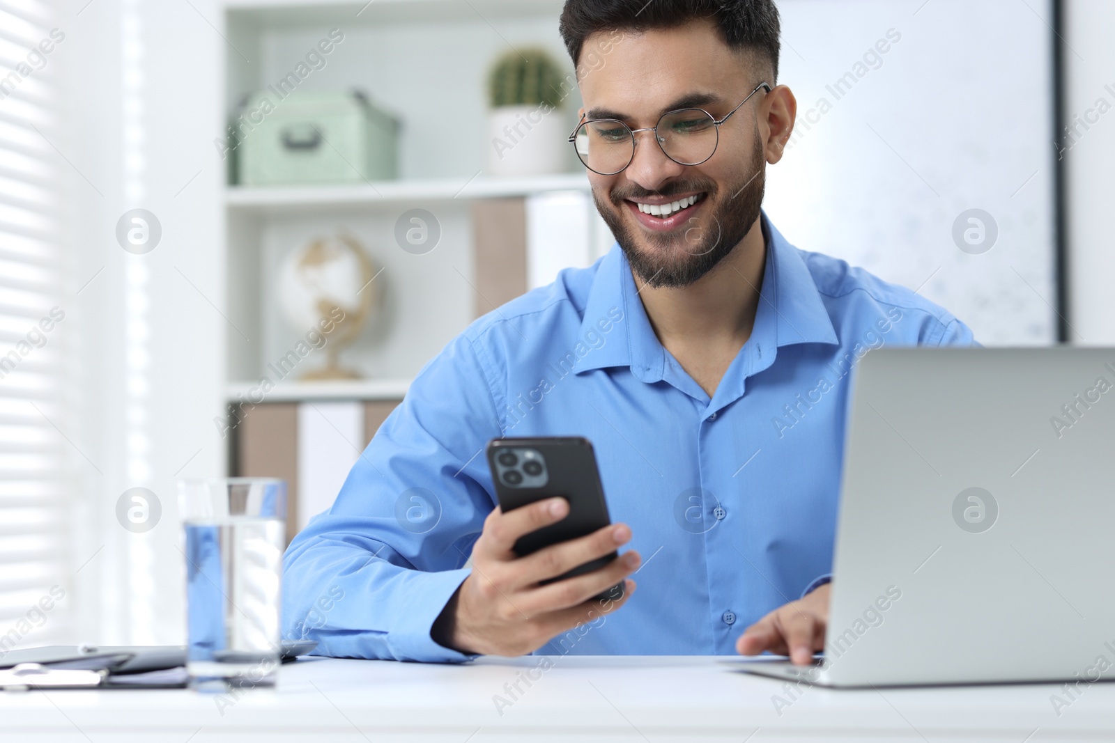 Photo of Happy young man using smartphone while working with laptop at white table in office
