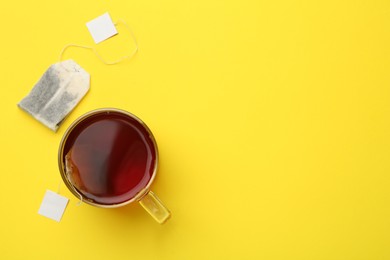 Photo of Tea bags and glass cup of hot drink on yellow background, flat lay. Space for text