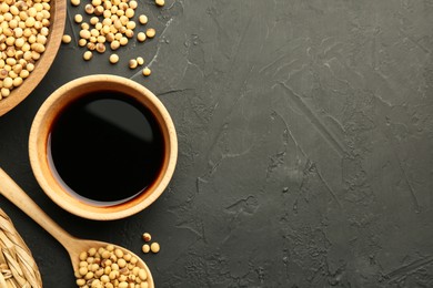 Photo of Soy sauce in bowl and soybeans on black textured table, flat lay. Space for text