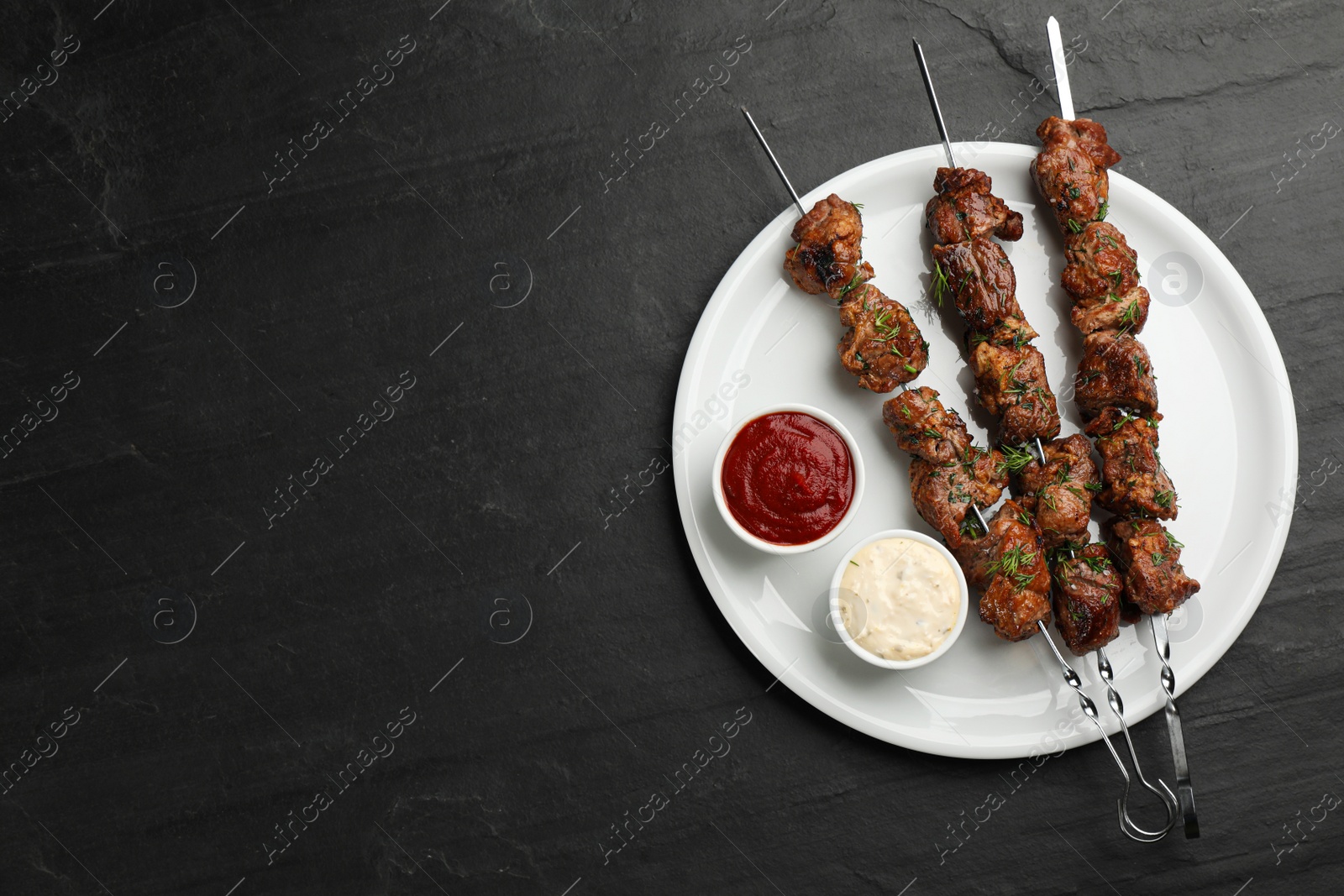 Photo of Metal skewers with delicious meat, ketchup and sauce on black table, top view. Space for text