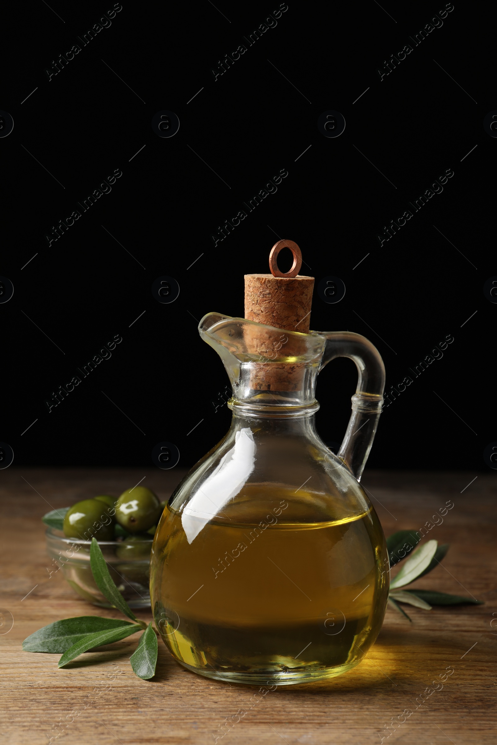 Photo of Glass jug of oil, ripe olives and green leaves on wooden table