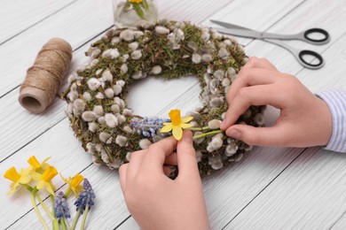 Photo of Woman decorating willow wreath with daffodil and hyacinth flowers at white wooden table, closeup