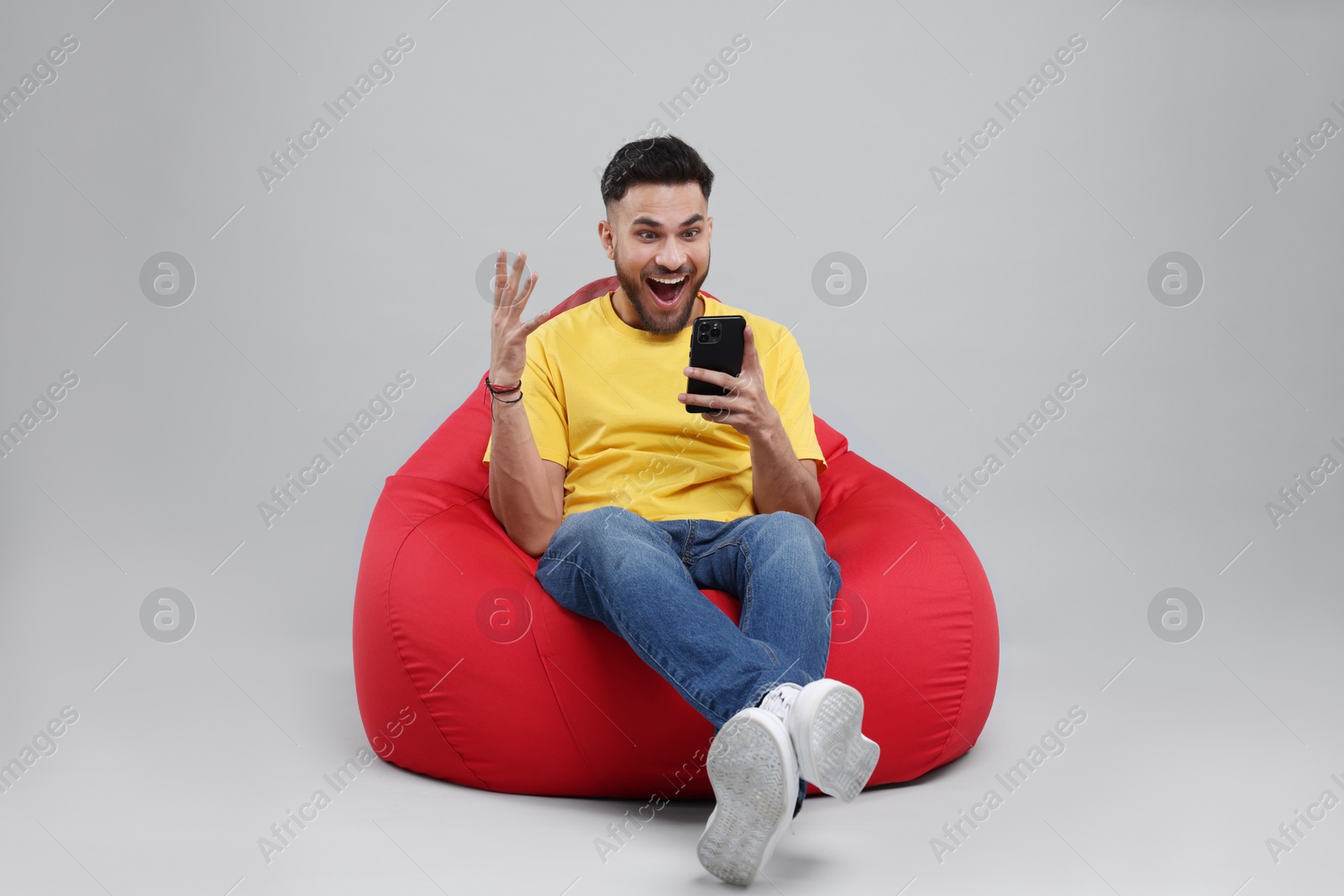 Photo of Emotional young man using smartphone on bean bag chair against grey background