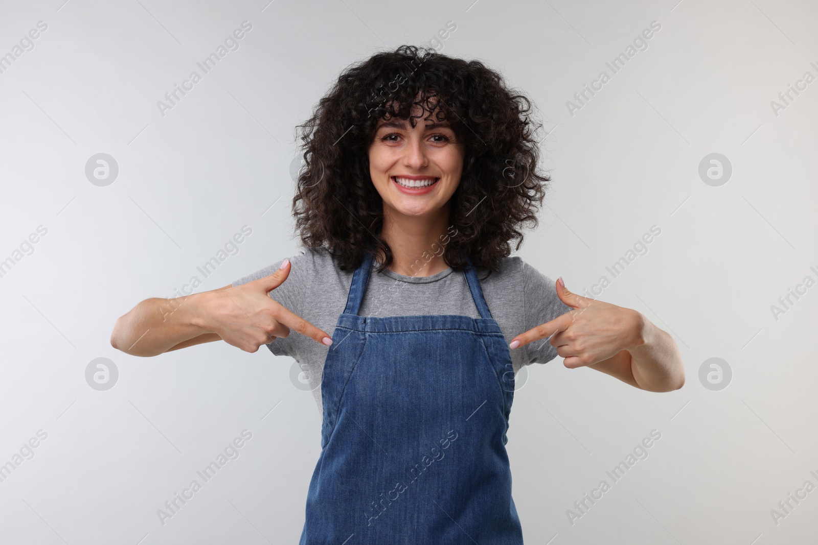 Photo of Happy woman pointing at kitchen apron on light grey background. Mockup for design