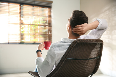 Young man with cup of drink relaxing near window at home, back view. Space for text