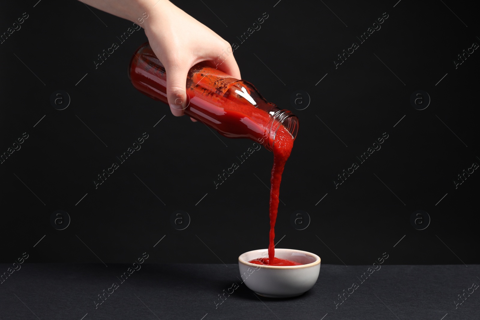 Photo of Woman pouring tasty ketchup from bottle into bowl against black background, closeup