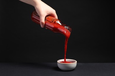 Woman pouring tasty ketchup from bottle into bowl against black background, closeup