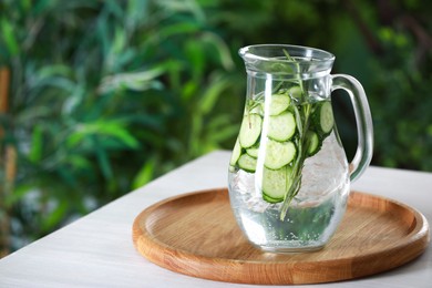 Refreshing cucumber water with rosemary in jug on white table against blurred green background. Space for text