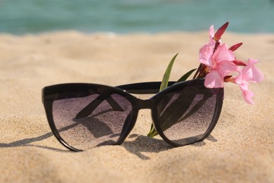 Photo of Beautiful sunglasses with flower on sand near sea, closeup