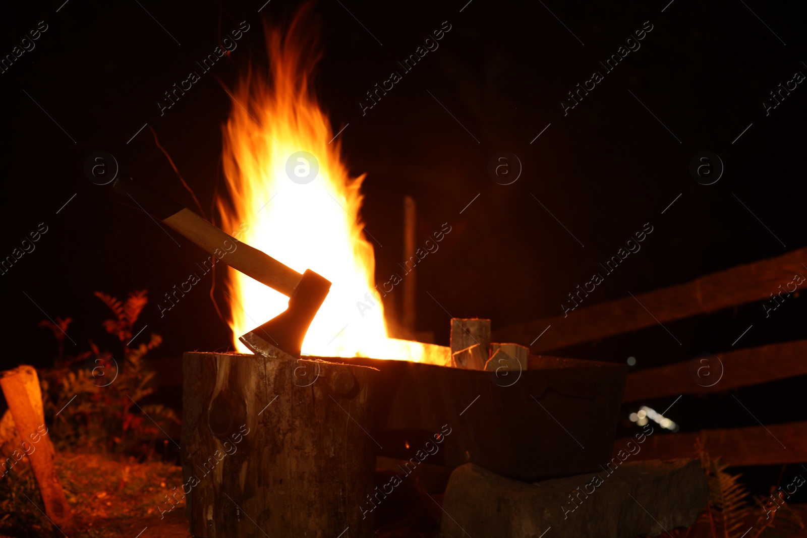 Photo of Tree stump with axe and burning firewood in metal brazier outdoors at night